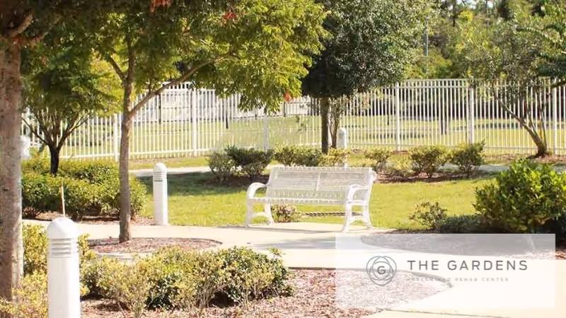 White metal bench on a paved path in a landscaped courtyard with trees, shrubs, and a fence in the background.