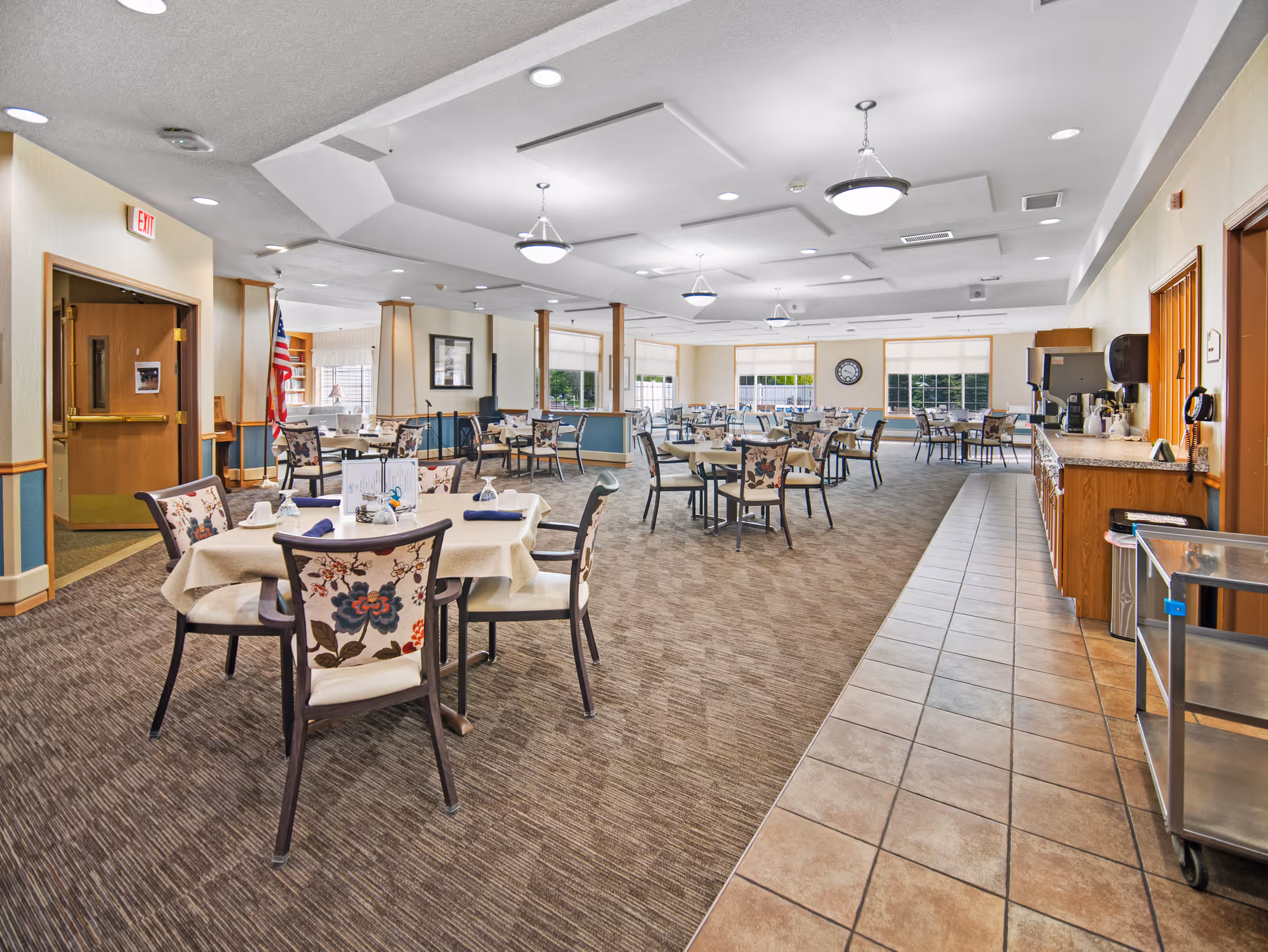 A spacious dining room in a senior living facility with multiple round tables covered with beige tablecloths and floral-patterned chairs. The room has carpeted flooring with a tiled walkway on the right side leading to a counter area with coffee machines and supplies. Large windows allow natural light to fill the room, and ceiling lights provide additional illumination. An American flag is visible near an open doorway on the left side.