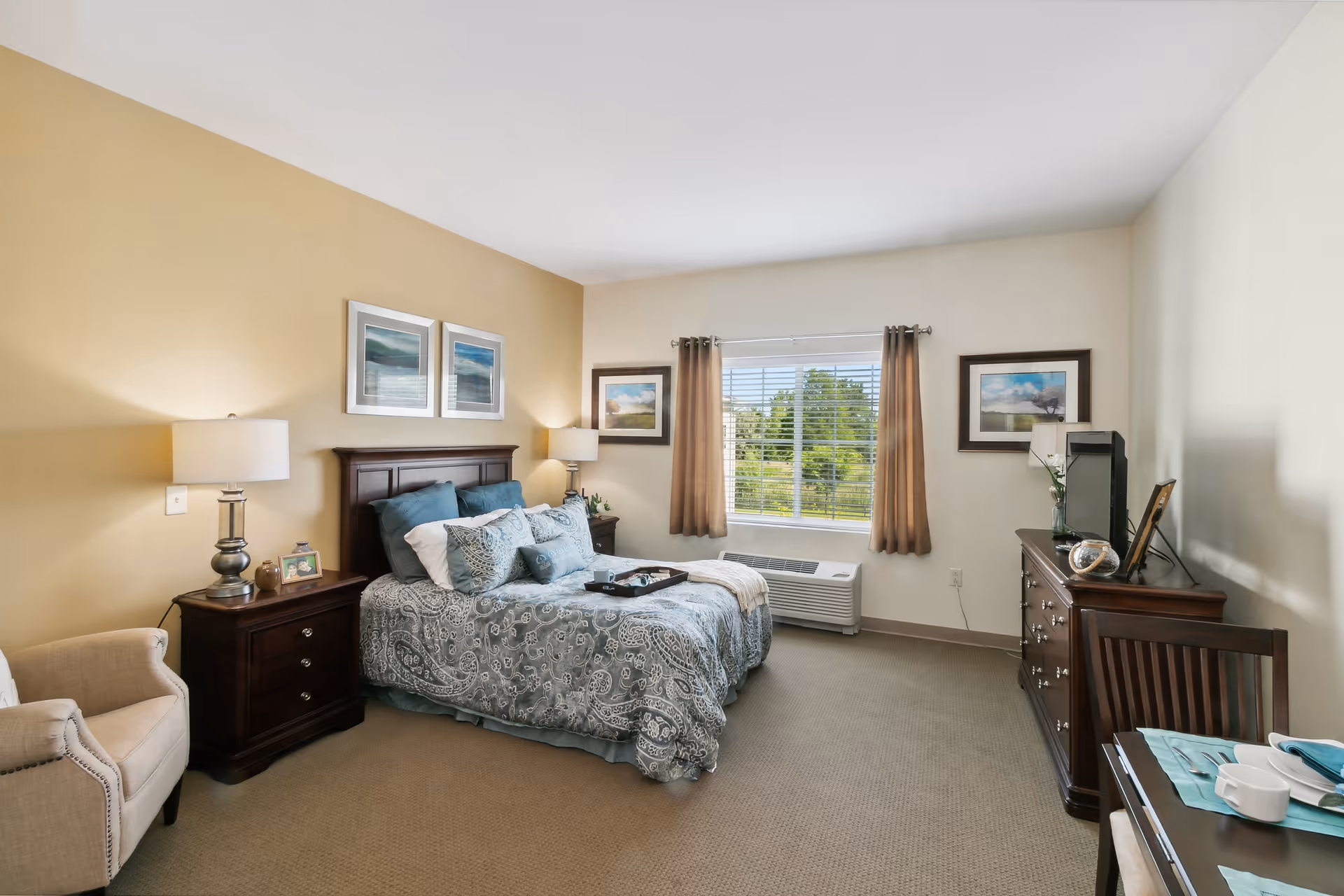 A well-lit bedroom with a large bed featuring blue and white patterned bedding and multiple pillows. On either side of the bed are dark wooden nightstands with lamps. There is a beige armchair to the left and a dark wooden dresser with a TV on top to the right. A window with brown curtains allows natural light to enter the room, and framed artwork hangs on the walls.