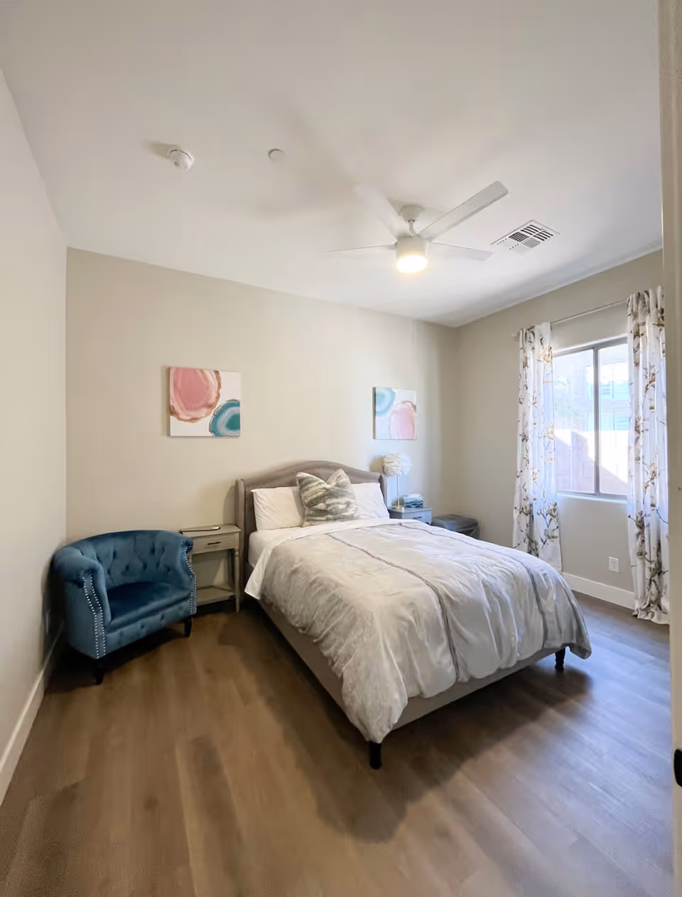 A bright bedroom with a neatly made bed featuring white and gray bedding, a blue upholstered armchair to the left, two small nightstands on either side of the bed, a window with floral curtains on the right wall, and two abstract paintings hanging above the bed. The room has light-colored walls, wood flooring, and a ceiling fan with a light.