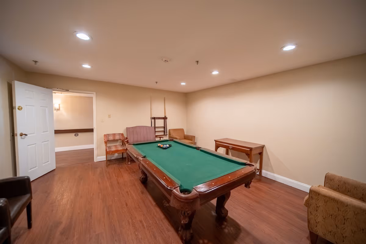 Interior room with a green felt pool table in the center, surrounded by several chairs and a small wooden table against the wall. The room has wooden flooring, beige walls, and recessed ceiling lights. An open door leads to another room with similar flooring and wall color.
