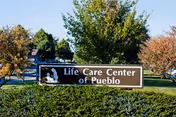 A brown sign reading "Life Care Center of Pueblo" sits on a grassy lawn with trees and a building visible behind it.