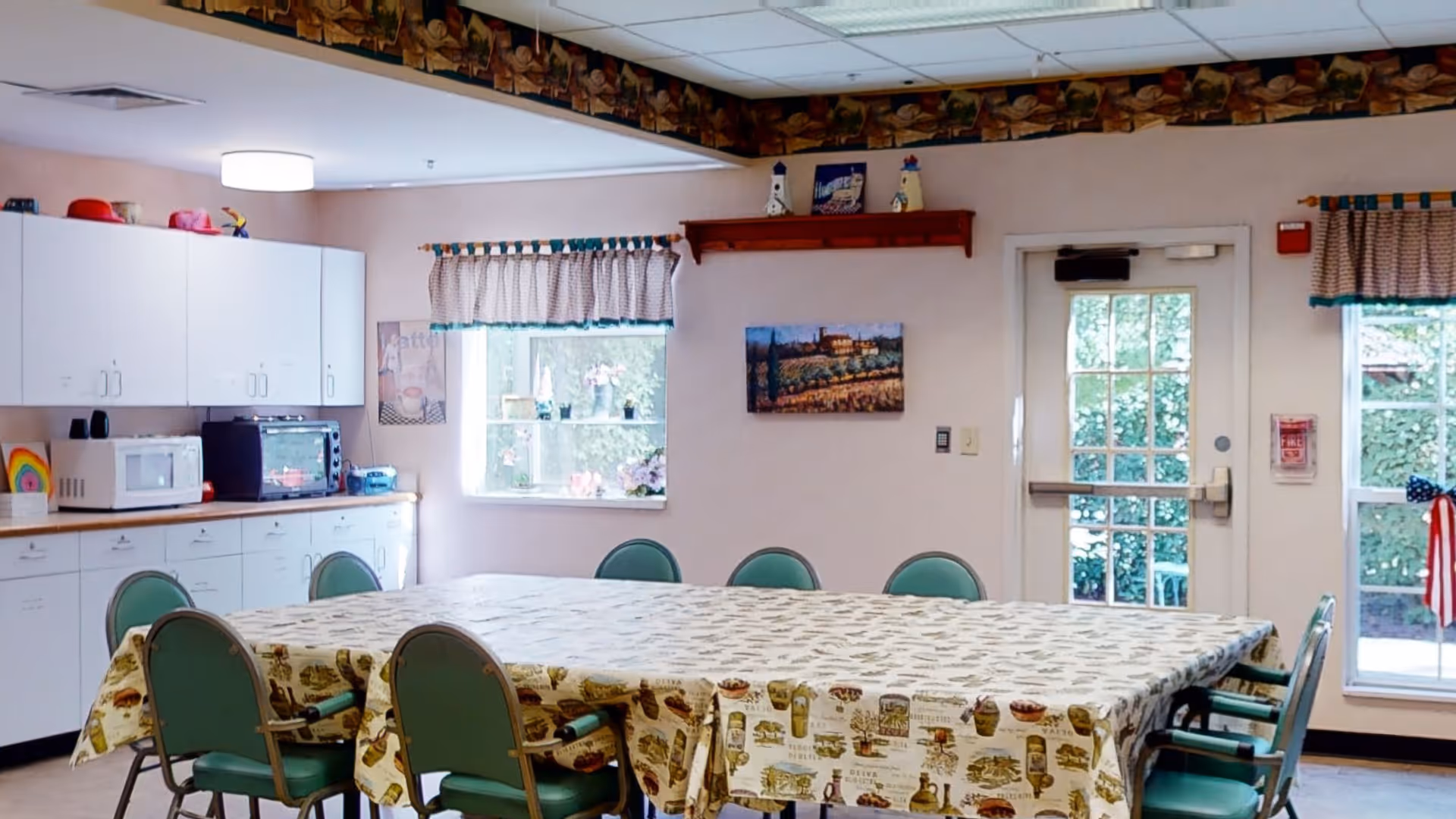 Communal dining/activity room with a large table covered in a patterned tablecloth, green chairs, cabinets, a microwave, and windows with valances.