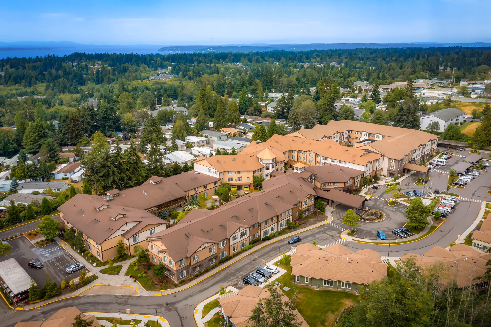 Aerial view of Quail Park of Lynnwood senior living facility showing multiple connected buildings with brown roofs surrounded by trees, parking lots, and roads, with a forested area and water body visible in the background.