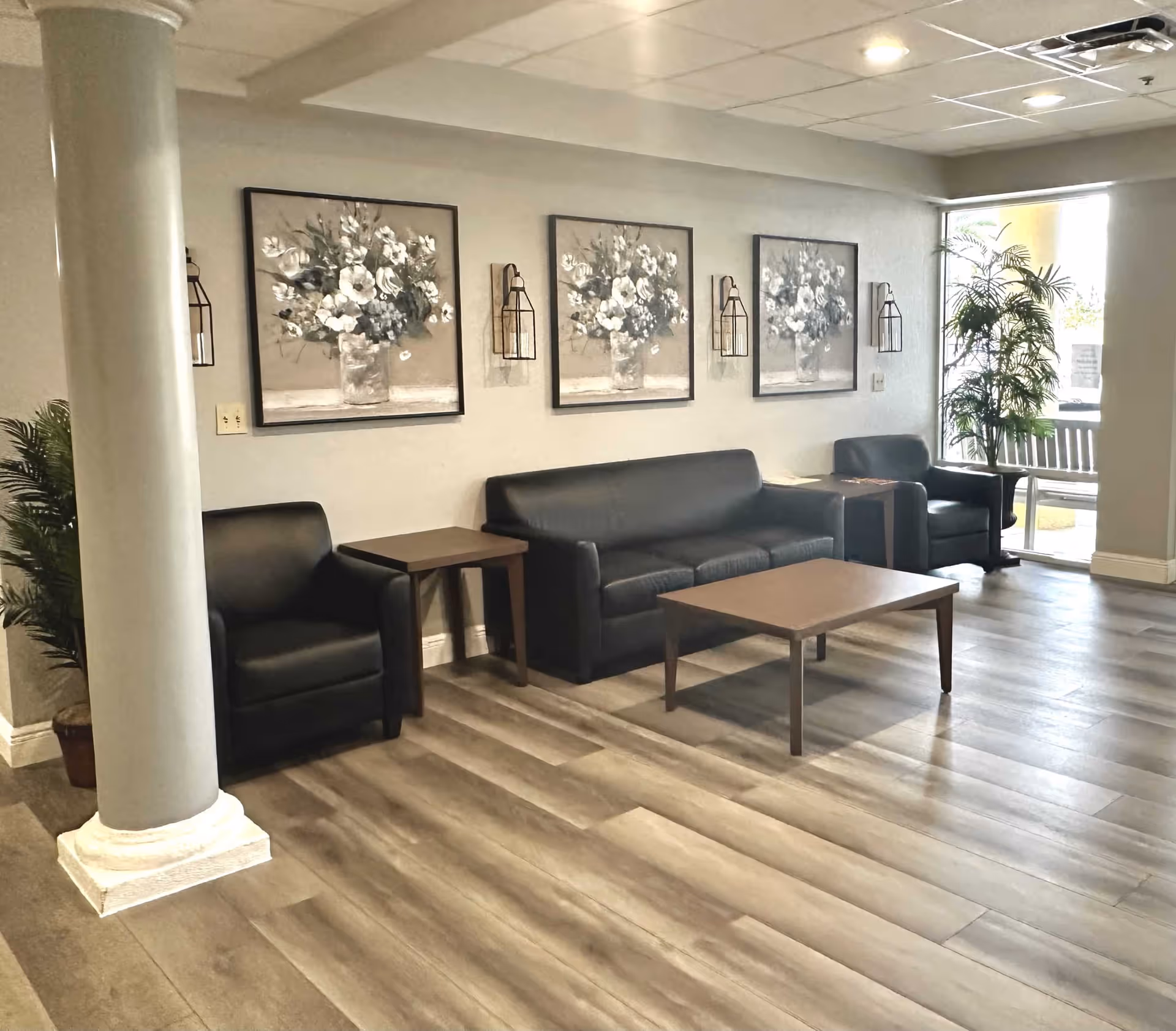 A seating area in a senior living facility with a black leather sofa, two black leather armchairs, two wooden side tables, and a wooden coffee table. The walls are decorated with three framed floral paintings and two wall-mounted lantern-style light fixtures. There is a large window with a potted plant next to it, and a column is visible on the left side of the image.