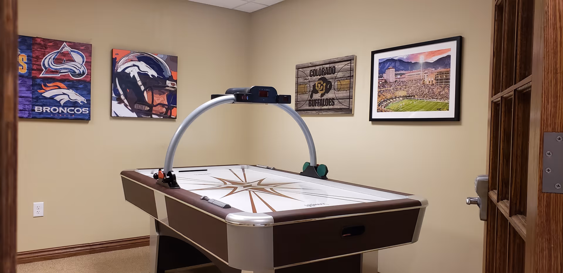 A room with an air hockey table in the center and four framed pictures on the beige walls, including sports-themed artwork featuring the Denver Broncos and Colorado Buffaloes. The room has a carpeted floor and a wooden door partially open on the right side.
