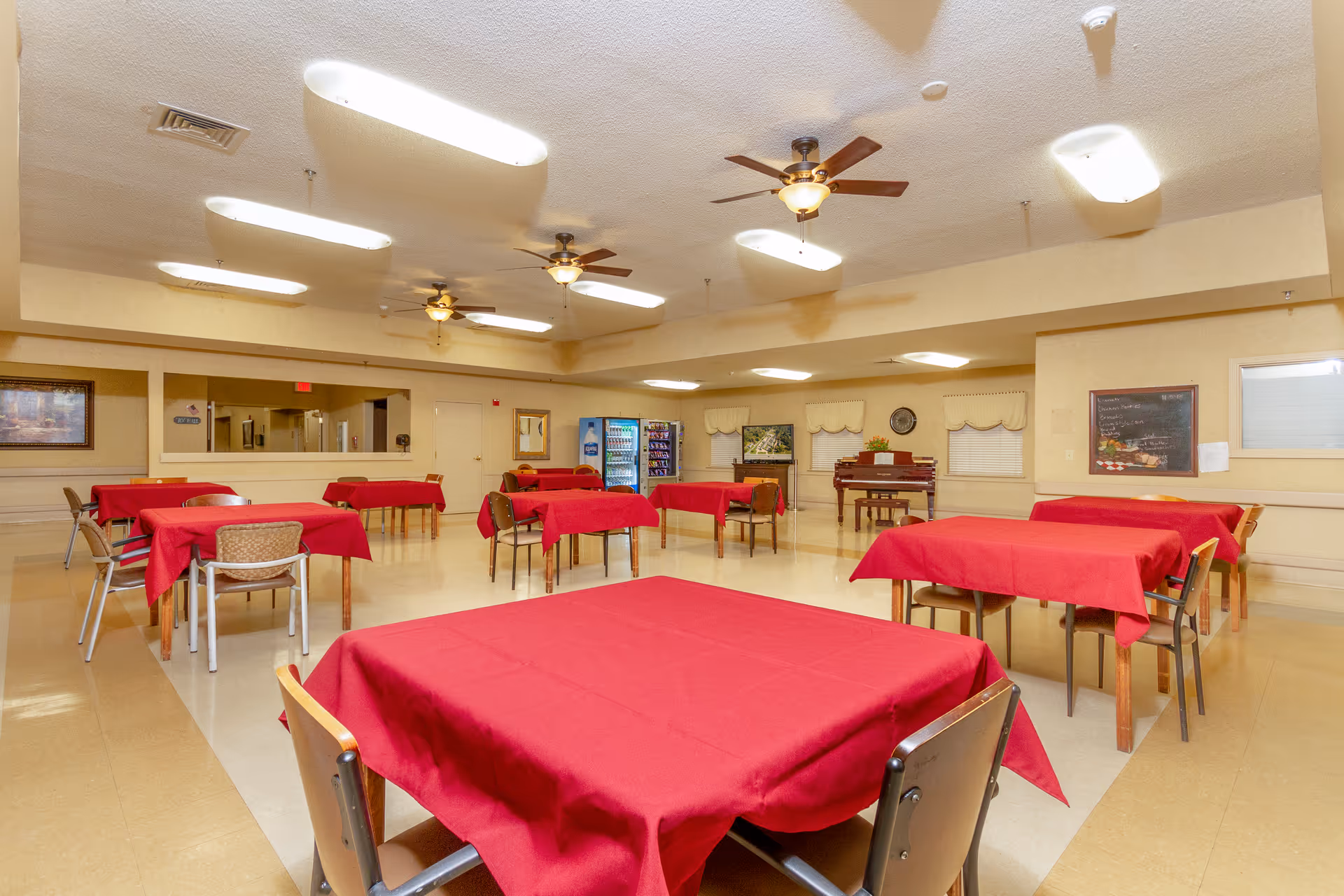 A spacious dining room with multiple tables covered in red tablecloths and surrounded by chairs. The room has beige walls, ceiling fans with lights, and fluorescent ceiling lights. There is a vending machine and a piano against the far wall, along with framed pictures and a chalkboard menu.