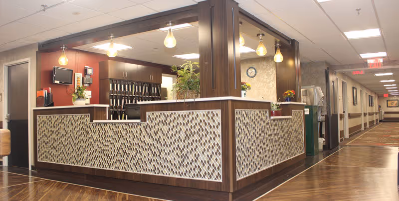 Reception desk area inside a senior living facility with a patterned front panel, hanging light bulbs, a small TV mounted on the wall, and a hallway extending to the right with framed pictures on the walls and exit signs visible.