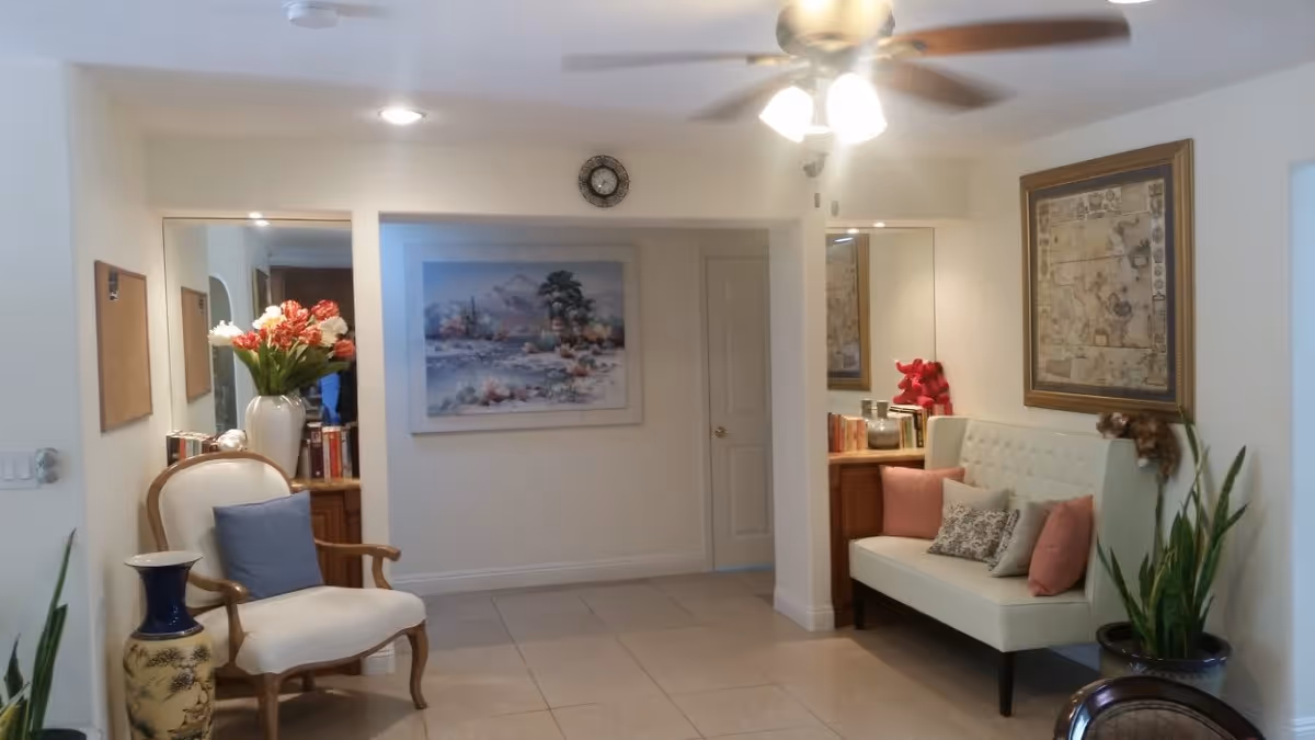 Well-lit seating area with an armchair, cushioned bench with pillows, artwork on the walls, plants, and a ceiling fan.