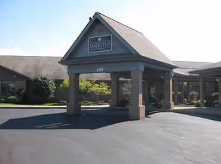 Entrance of a single-story building with a covered driveway supported by brick columns. The building has a sign that reads 'Emeritus' above the entrance and the number 100 is displayed on the structure. There are shrubs and small trees around the building under a clear sky.