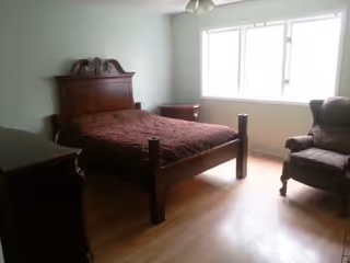 Bedroom with a wooden bedframe, dresser, armchair and a large window letting in natural light.