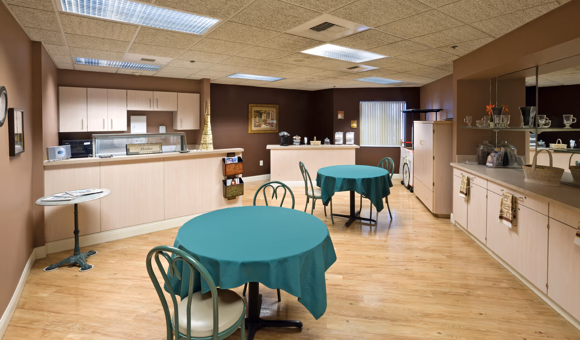 Interior view of a dining area in a senior living facility with two round tables covered with green tablecloths and green chairs. The room has light wood flooring, brown walls, and a ceiling with fluorescent lights. There is a counter with cabinets and a small service area in the background, along with a sideboard with glassware and decorative items on the right side.