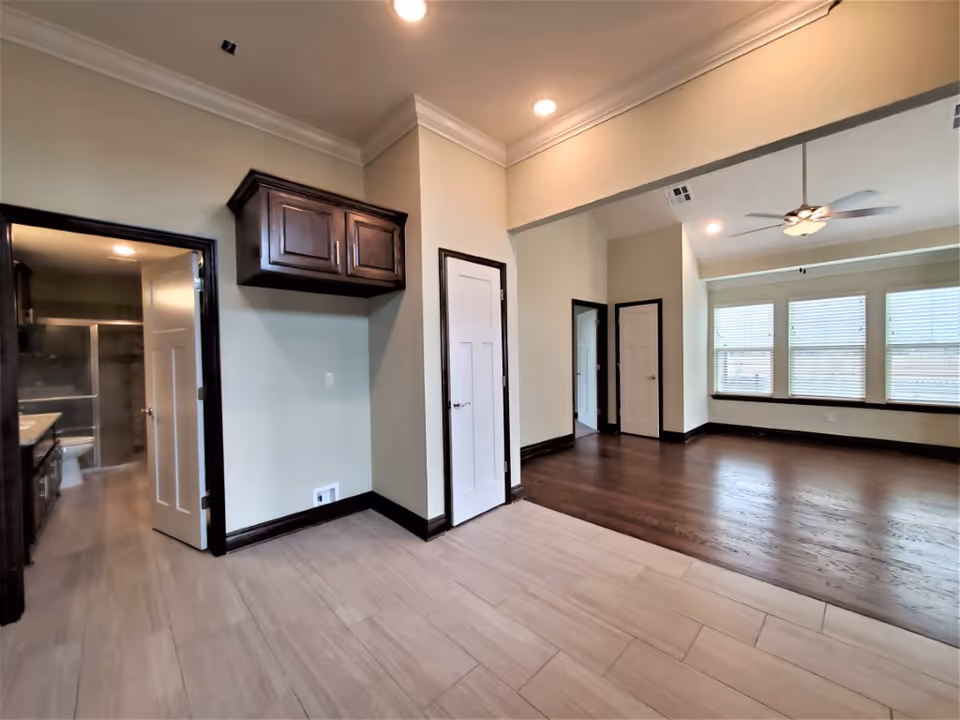 Spacious open interior showing tile and hardwood floors, large windows with blinds, a ceiling fan, and several doors to adjoining rooms.
