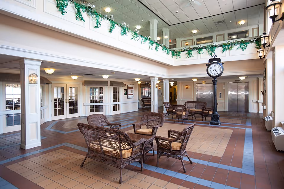 Spacious and well-lit common area in a senior living facility with wicker chairs and sofas arranged around small tables. The room features a tiled floor, white pillars, a large black standing clock, and two elevators in the background. Green plants decorate the upper balcony railing.