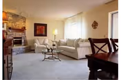 Bright living room with a striped sofa, armchair, glass coffee table, stone fireplace, and dining table visible in the foreground.