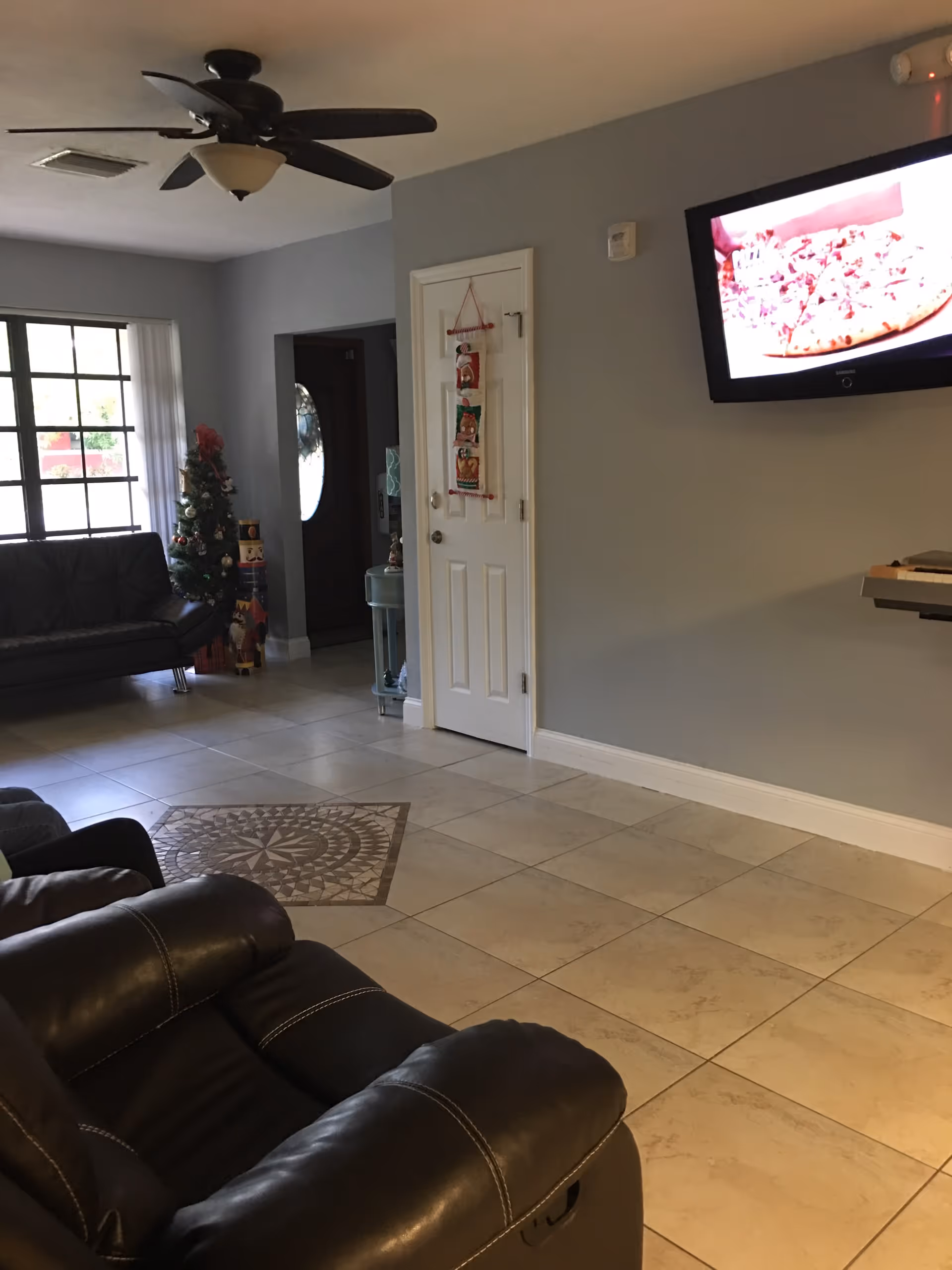 Interior view of a living room with beige tiled floor and a decorative tile pattern in the center. There are two black leather recliners in the foreground and a black leather couch near a window with vertical blinds. A small decorated Christmas tree and holiday decorations are visible near the window. A ceiling fan with lights is mounted on the ceiling. A white door with hanging holiday decorations is on the right wall, and a flat-screen TV mounted on the wall displays an image of a pizza.