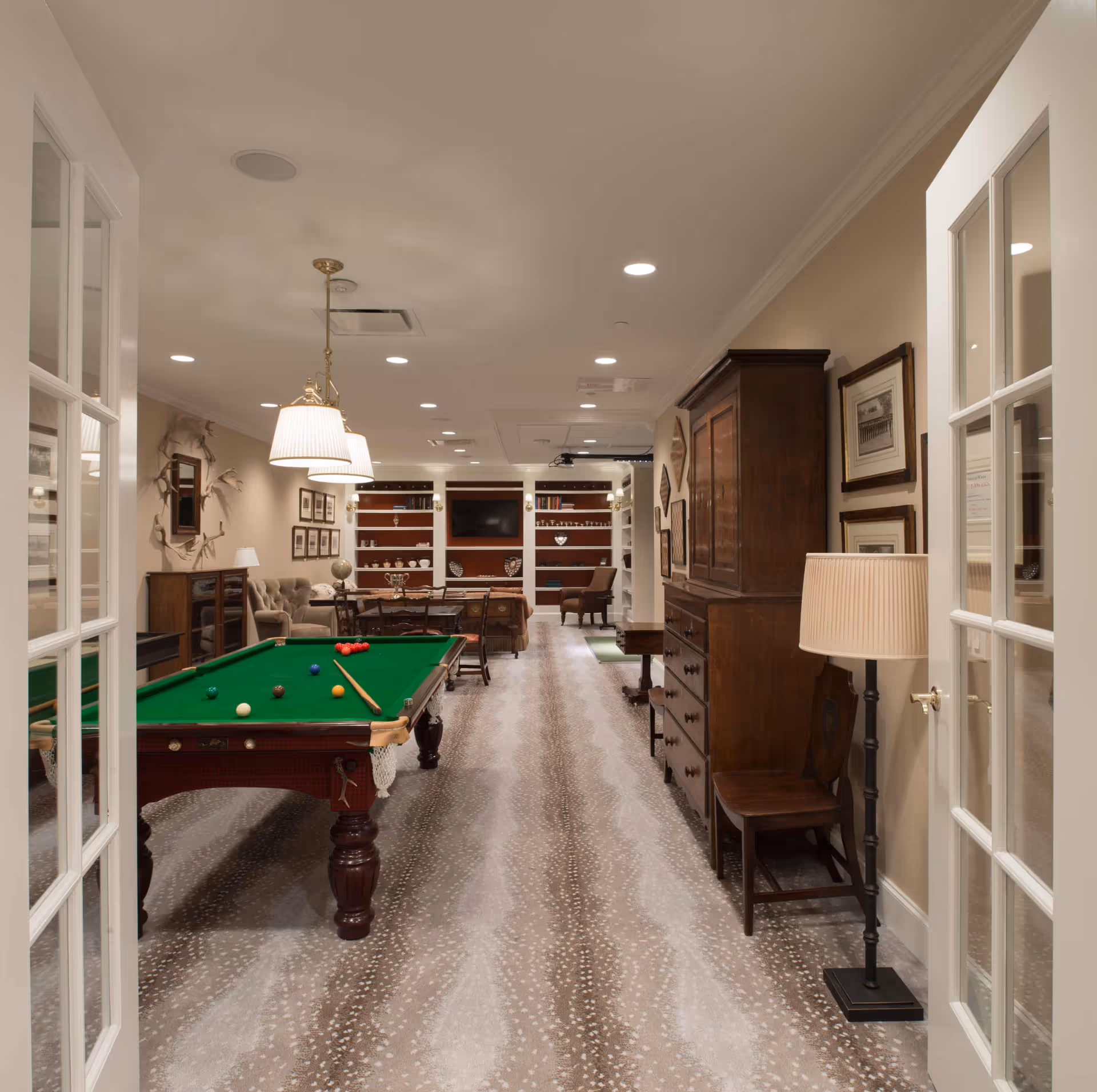 Interior view of a recreational room with a pool table in the foreground, a wooden cabinet and chair on the right, and a seating area with a sofa, chairs, and a TV on the far wall. The room has patterned carpet flooring, recessed ceiling lights, and pendant lamps above the pool table.
