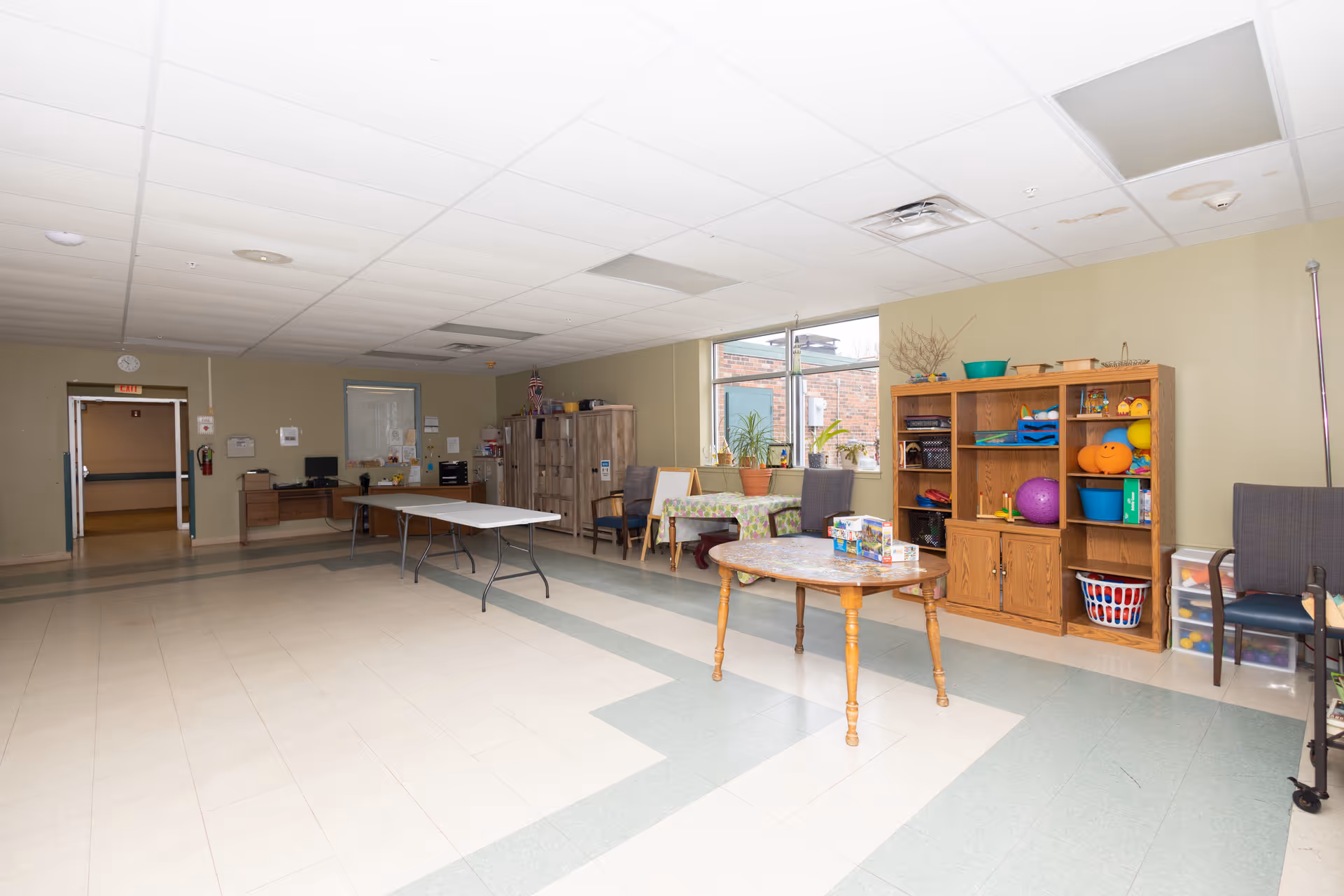 Spacious multipurpose activity room with folding tables, a round table, shelving filled with games and exercise balls, chairs, and large windows.