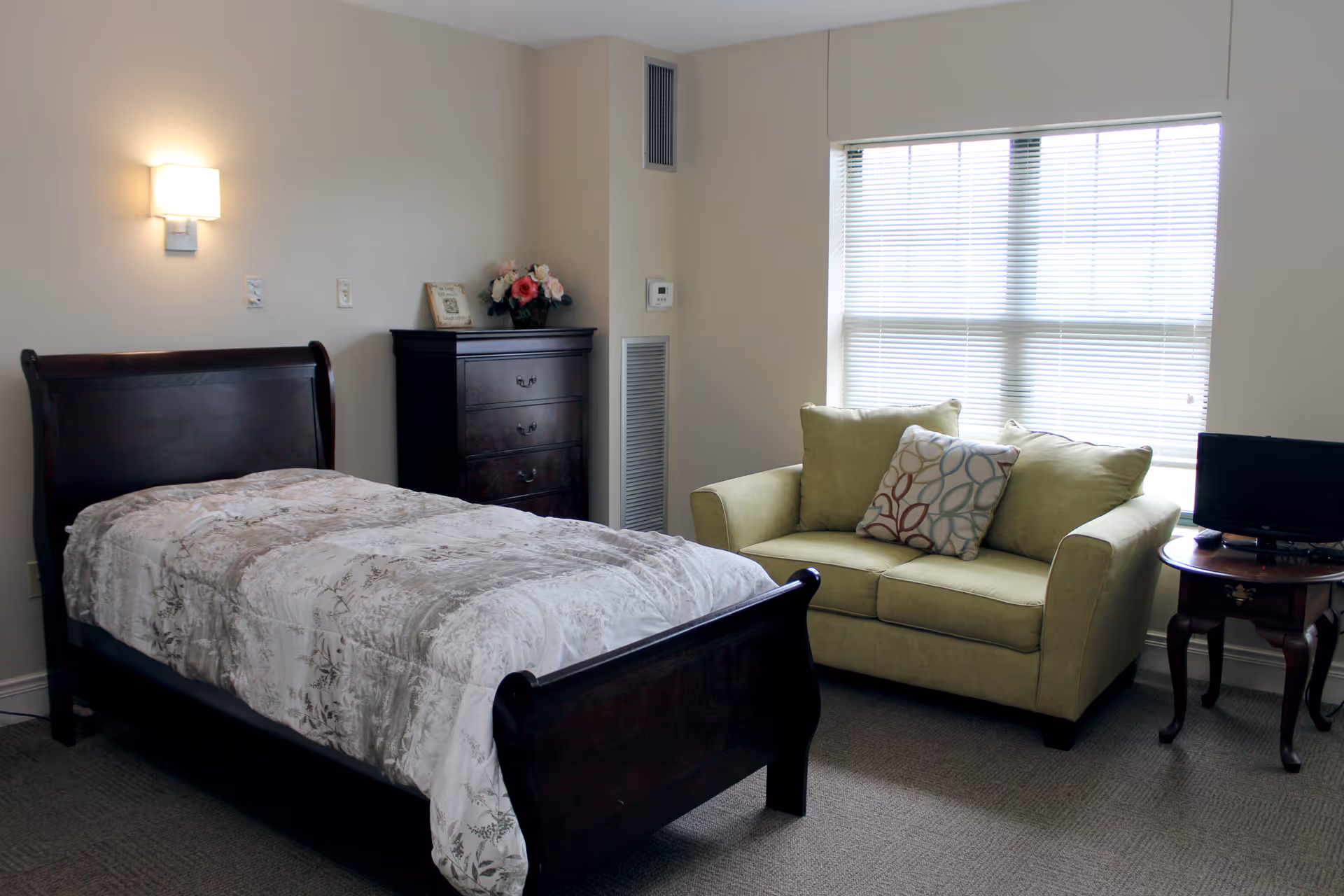 A cozy bedroom in an assisted living facility featuring a dark wooden single bed with a patterned comforter, a matching dark wooden chest of drawers with a flower arrangement and picture frame on top, a green loveseat with two pillows, a small wooden side table with a small TV, and a large window with blinds letting in natural light.