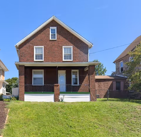 Front exterior view of a two-story brick house with a small covered porch, white door, and several windows, situated on a grassy lawn under a clear blue sky.