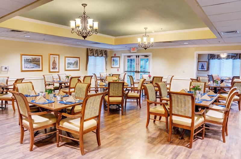 A bright and spacious dining room with multiple wooden tables and chairs arranged neatly. Each table is set with folded napkins, glasses, and small flower arrangements. The room features wooden flooring, framed artwork on the walls, large windows with curtains, and elegant chandeliers hanging from the ceiling.