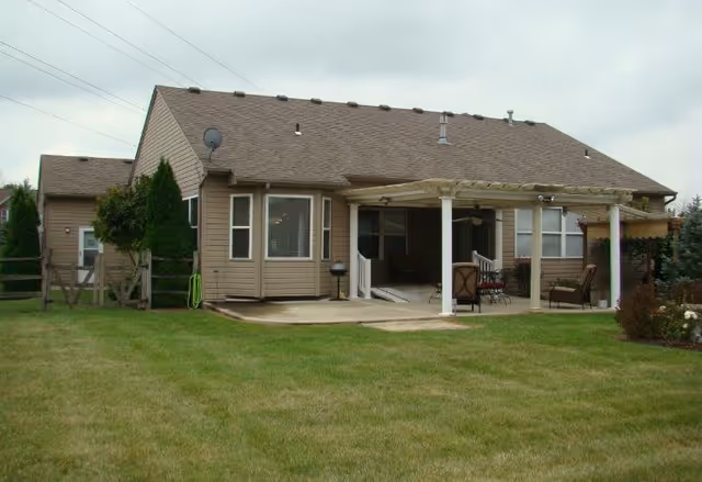 Back exterior view of a single-story house with beige siding and a brown shingled roof. The house features a covered patio area with outdoor furniture and a small grill. The yard is grassy and well-maintained with some shrubs and a wooden fence on the left side.