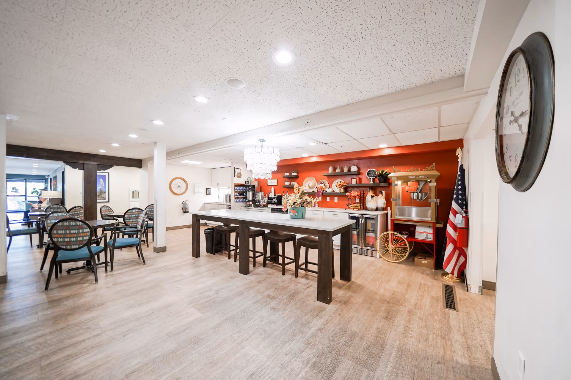 A bright and spacious dining area in a senior living facility featuring a large central table with stools, several smaller tables with chairs, a red accent wall with shelves displaying decorative items, a popcorn machine, and an American flag in the corner. The room has light wood flooring, recessed ceiling lights, and a large wall clock.