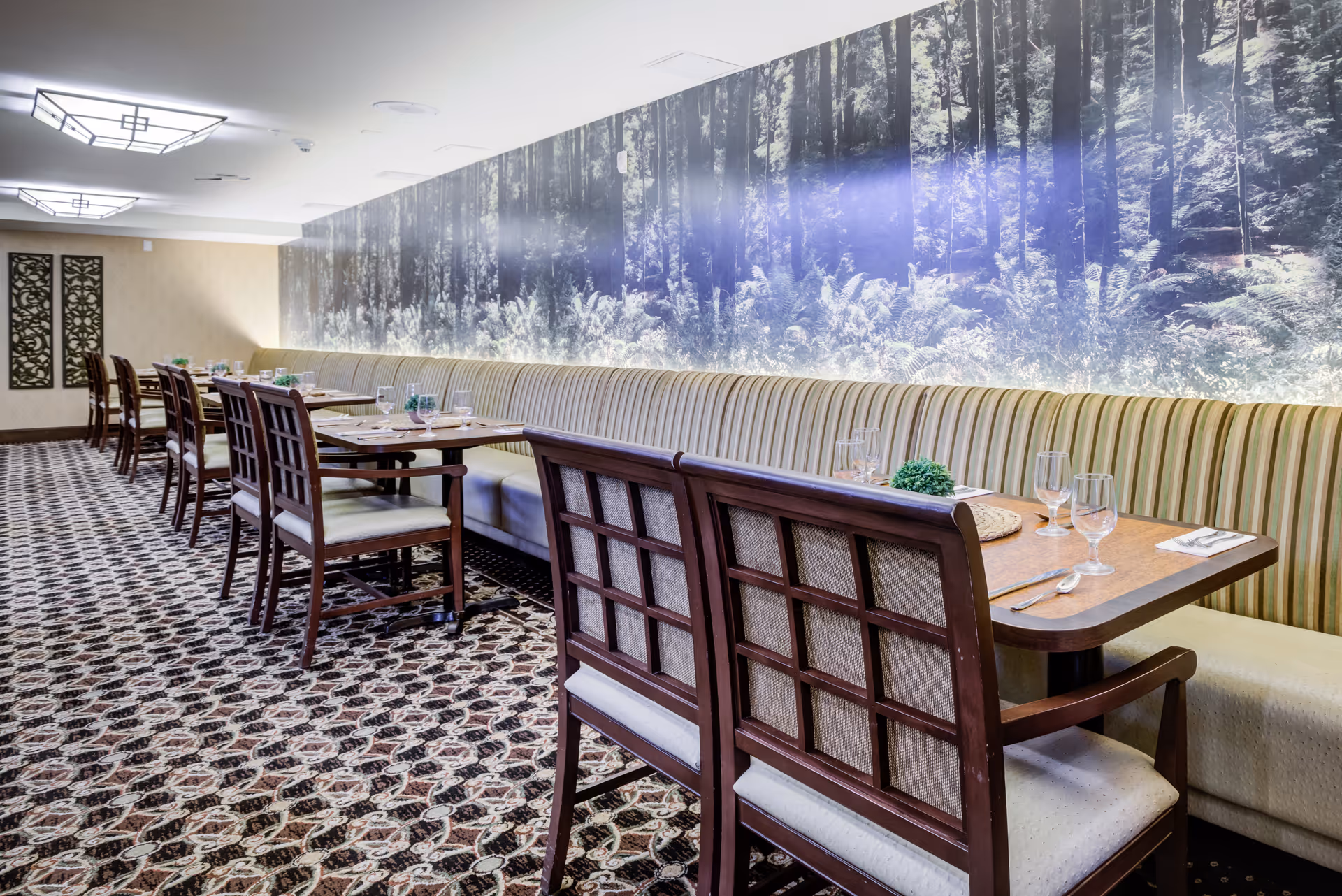 Dining room with a long banquette, wooden tables and chairs set with place settings, patterned carpet, and a large forest mural on the wall.