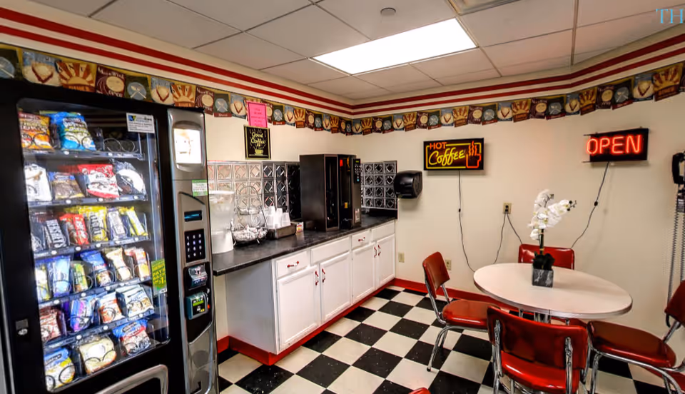 Small snack area with a vending machine, coffee station, countertop cabinets, and a round table with red chairs on a black-and-white checkered floor.