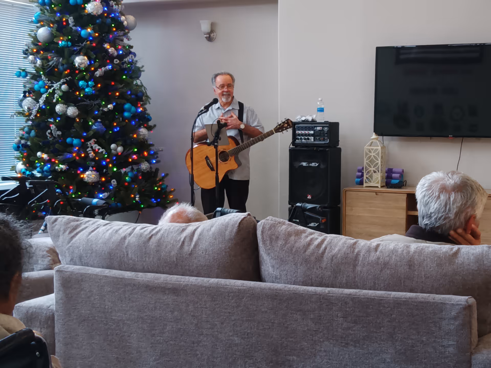 A man playing an acoustic guitar and singing into a microphone in a living room decorated with a large Christmas tree with colorful lights. Several elderly people are seated on couches facing him. A TV is mounted on the wall and a speaker system is set up nearby.