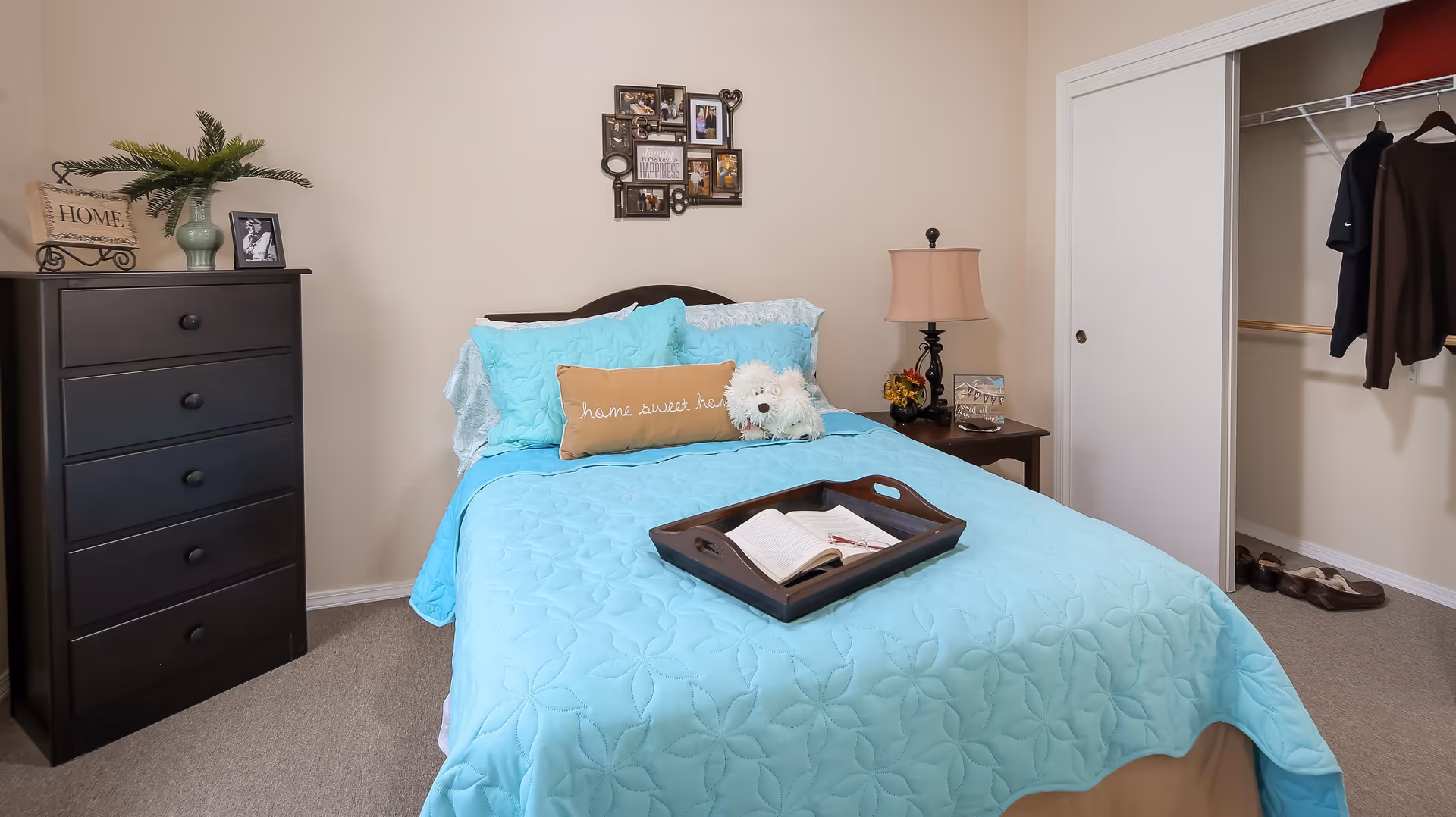 A cozy bedroom with a bed covered in a light blue quilt and pillows, including a decorative pillow that says 'home sweet home' and a small white stuffed dog. There is a dark wooden dresser with a plant and framed photo on top to the left, a nightstand with a lamp and decorative items to the right, and an open closet with clothes and shoes visible.