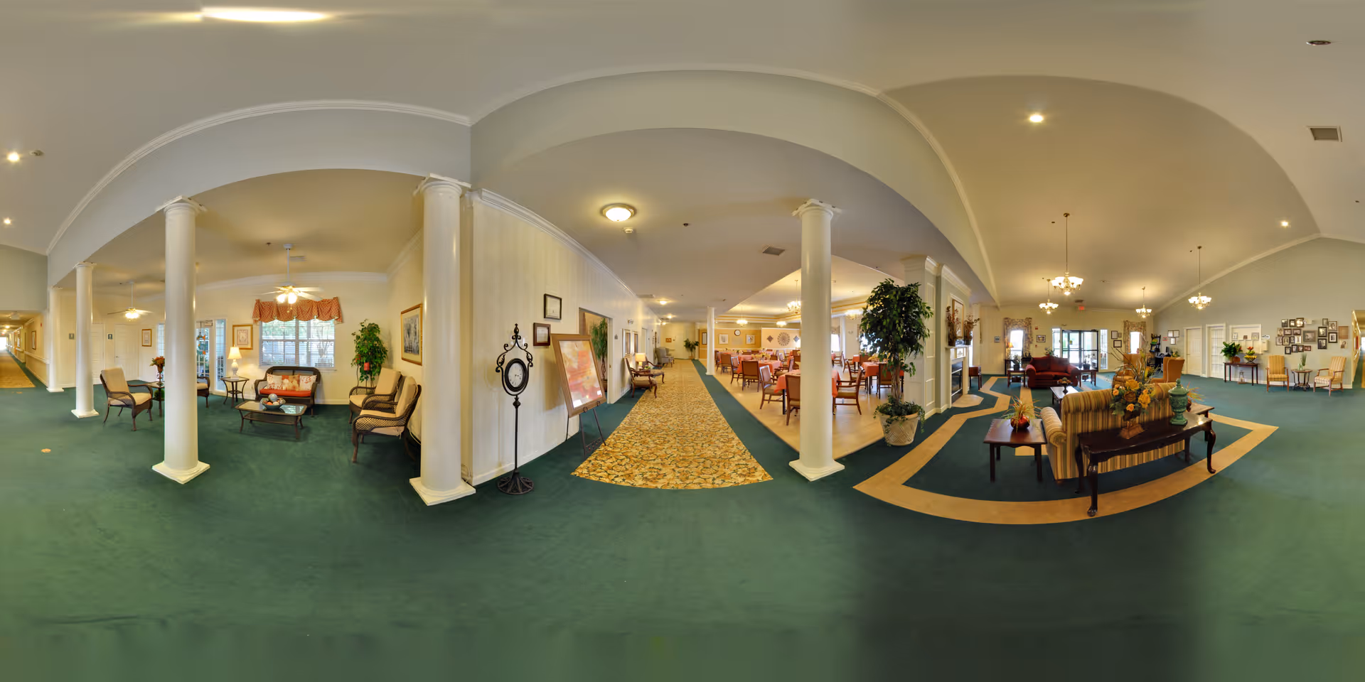 Spacious furnished senior living facility common area with seating, dining tables, columns, and green carpeting.