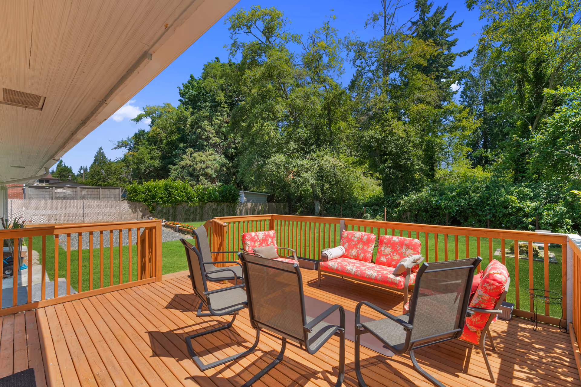 A wooden outdoor deck with a seating area consisting of a floral-patterned cushioned sofa and chair, along with four black mesh chairs around a rectangular table. The deck overlooks a green lawn and is surrounded by tall trees and bushes under a clear blue sky.