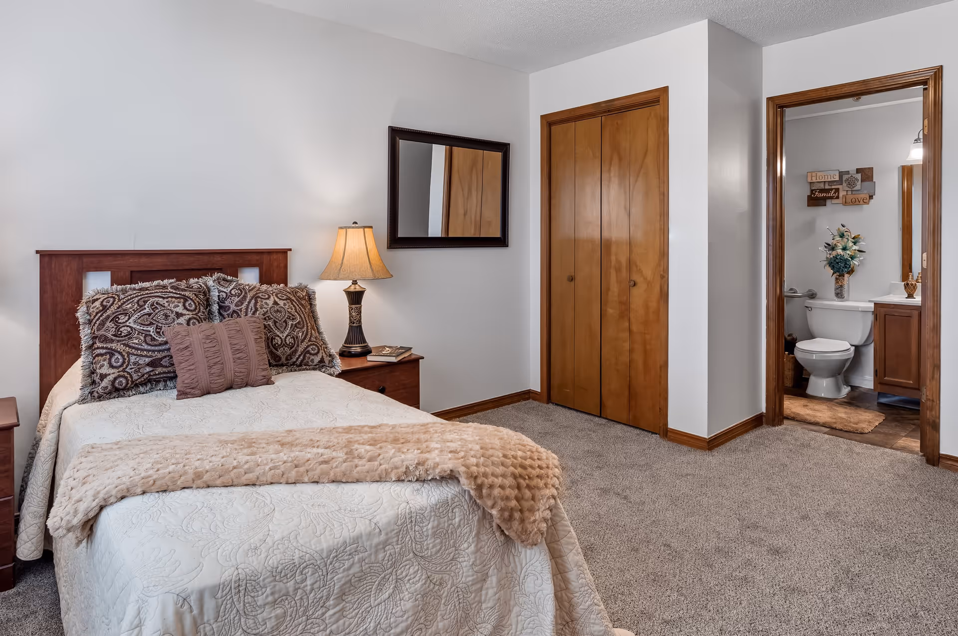 A cozy bedroom with a single bed covered in a white quilt and decorative pillows. A wooden nightstand with a lamp and books is beside the bed. There is a large mirror on the wall above the nightstand. The room has beige carpet and wooden trim. A wooden closet with double doors is visible, and an open doorway leads to a bathroom with a toilet, vanity, and floral arrangement.