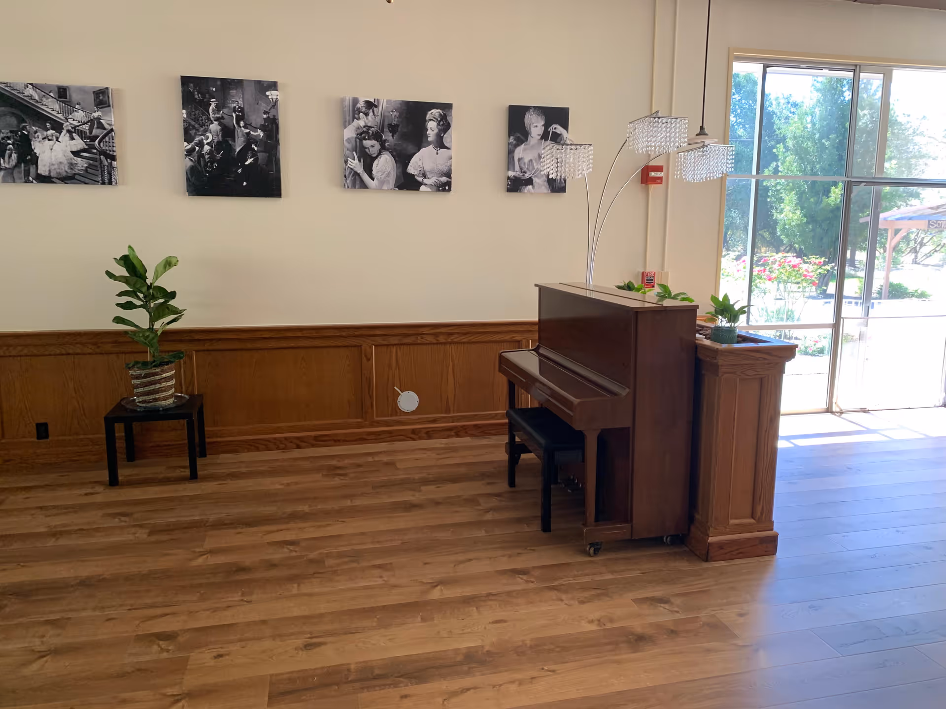 Interior view of a room with wooden flooring and wood paneling on the lower half of the walls. A dark wooden upright piano with a bench is positioned near a large window letting in natural light. On top of the piano is a modern lamp with three crystal-like shades and some small potted plants. On the left side of the room, there is a small black table with a potted plant. Four black and white framed photographs are hung on the beige wall above the wood paneling.