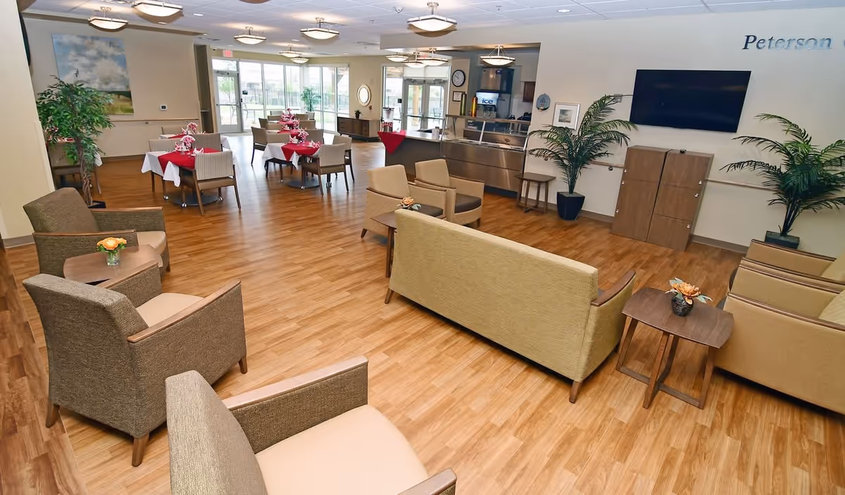 Spacious senior living community dining and lounge area with tables set with red napkins, sofas, chairs, plants, and a wall-mounted TV.
