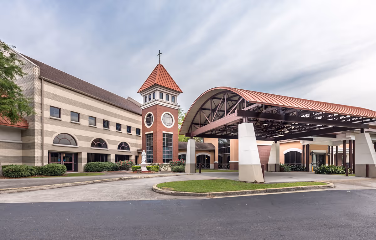 Front entrance of a senior living facility with a covered drive-through canopy, a brick tower topped with a cross, and arched windows.