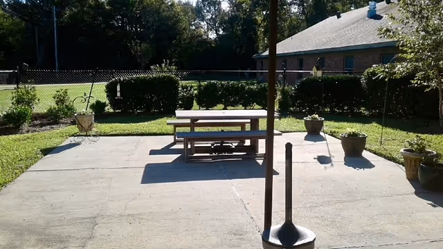 Concrete patio with a wooden picnic table, potted plants, a hedge, and a building in the background.