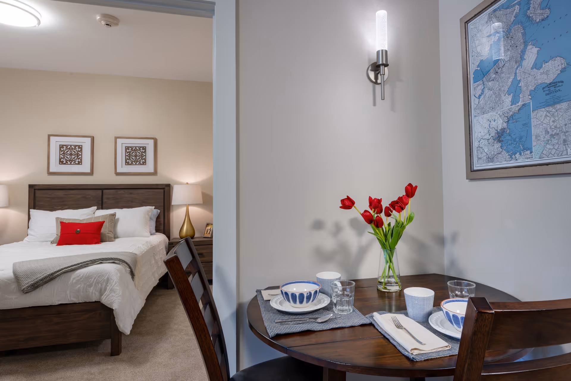 A cozy interior view showing a small dining table set for two with bowls, cups, glasses, and a vase of red tulips. To the left, there is a bedroom with a wooden bed frame, white bedding, a red accent pillow, two bedside lamps, and framed artwork on the wall.