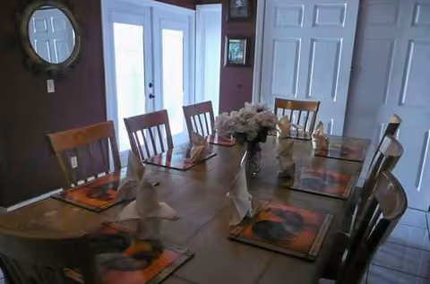 A dining room table is set with folded napkins, flowers, and placemats.