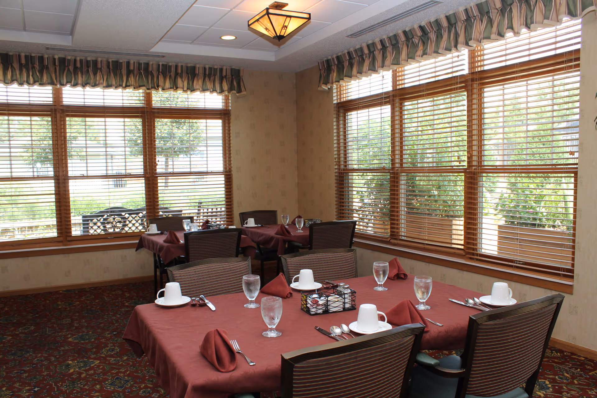 Dining room with tables covered in maroon tablecloths, set with white cups, glasses of water, silverware, and folded maroon napkins. Large windows with wooden blinds allow natural light to fill the room, and patterned carpet covers the floor.