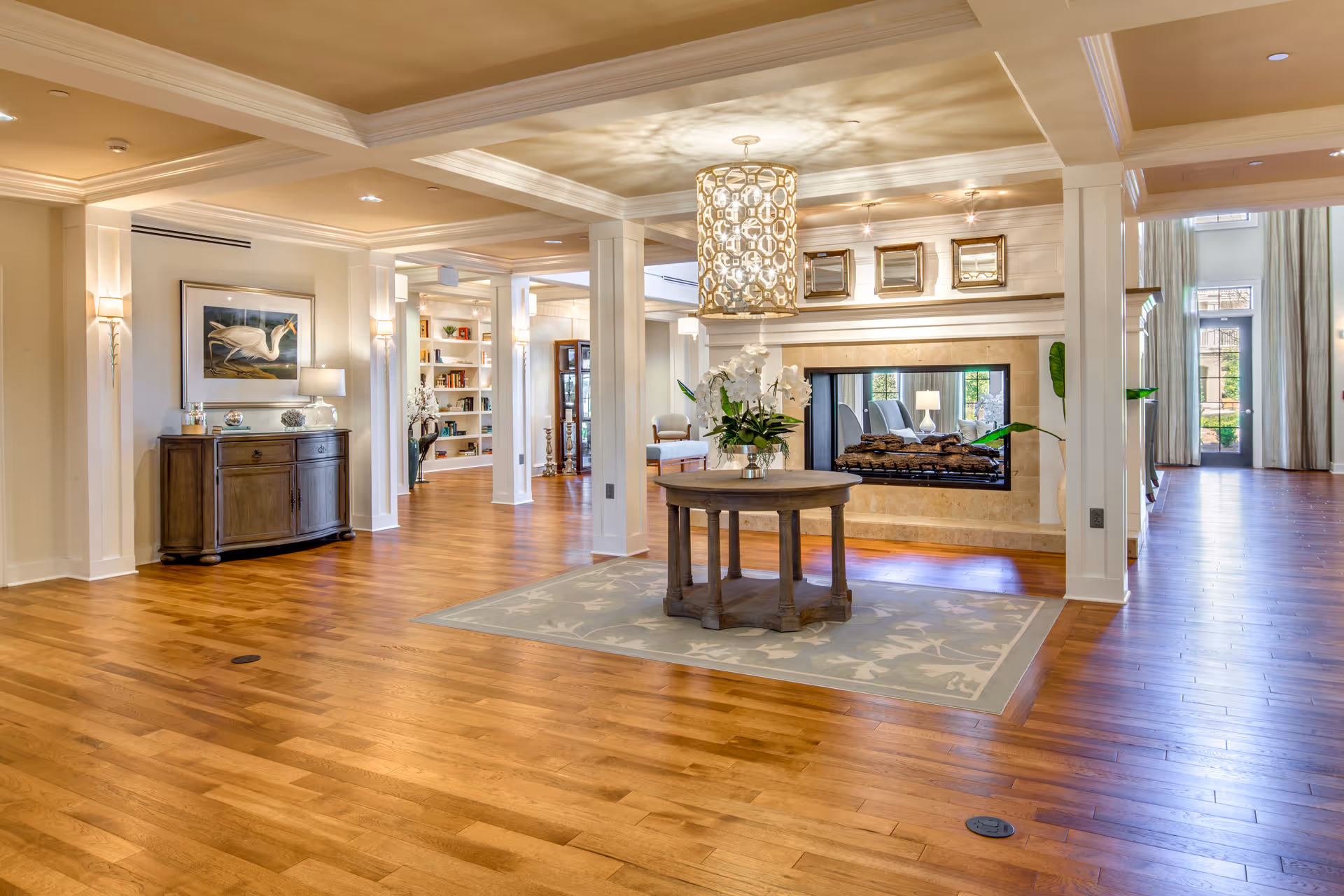 Spacious and well-lit interior common area with wooden flooring, a round table with a floral arrangement on a rug, a modern fireplace, decorative ceiling light fixture, and built-in shelves in the background.