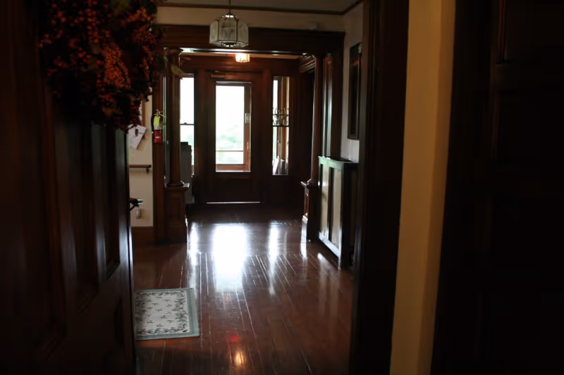 View down a dimly lit hallway with polished wooden floors and dark wood trim. At the end of the hallway is a wooden door with glass panels letting in natural light. A small rug is placed on the floor near the left side, and a decorative wreath hangs on the left wall. A radiator cover is visible on the right side of the hallway.