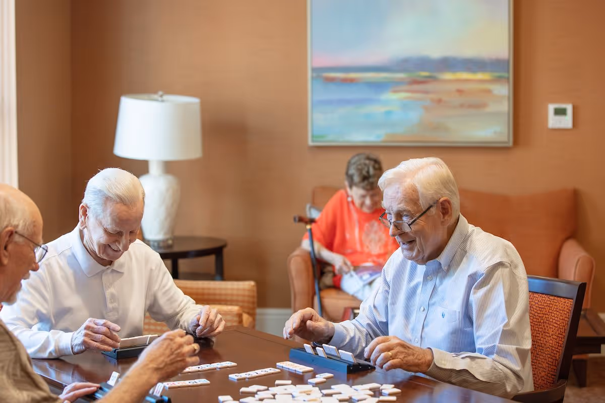 Three elderly men sitting around a wooden table playing a tile-based game, smiling and engaged. In the background, an elderly woman in an orange shirt is seated on an orange couch, reading a book. The room has warm brown walls, a white table lamp on a side table, and a colorful landscape painting on the wall.