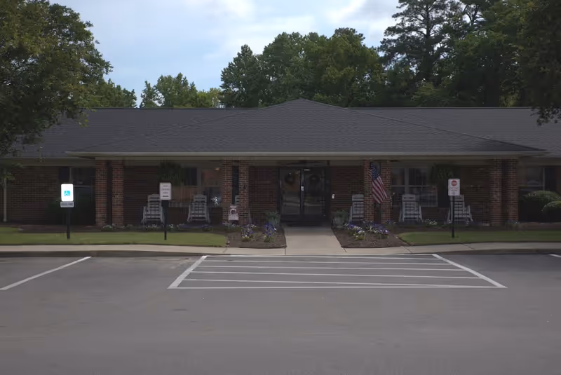 Front exterior view of a single-story brick building with a covered entrance, several rocking chairs on the porch, an American flag, and parking spaces including a handicapped spot in front.