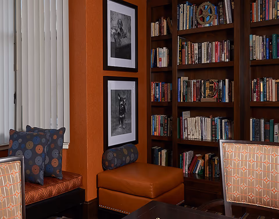 Cozy interior corner of a room with a built-in wooden bookshelf filled with books, two framed black and white dog portraits on an orange wall, a cushioned bench with patterned pillows, and a leather ottoman.