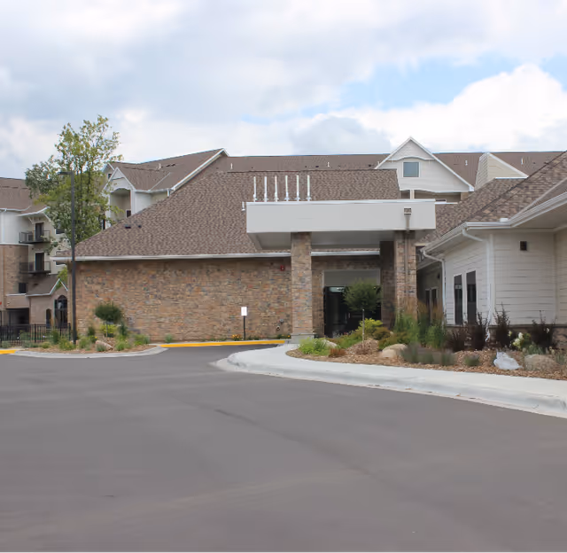 Exterior view of a senior living facility building with a covered entrance, stone and siding walls, landscaped plants, and a paved driveway under a partly cloudy sky.