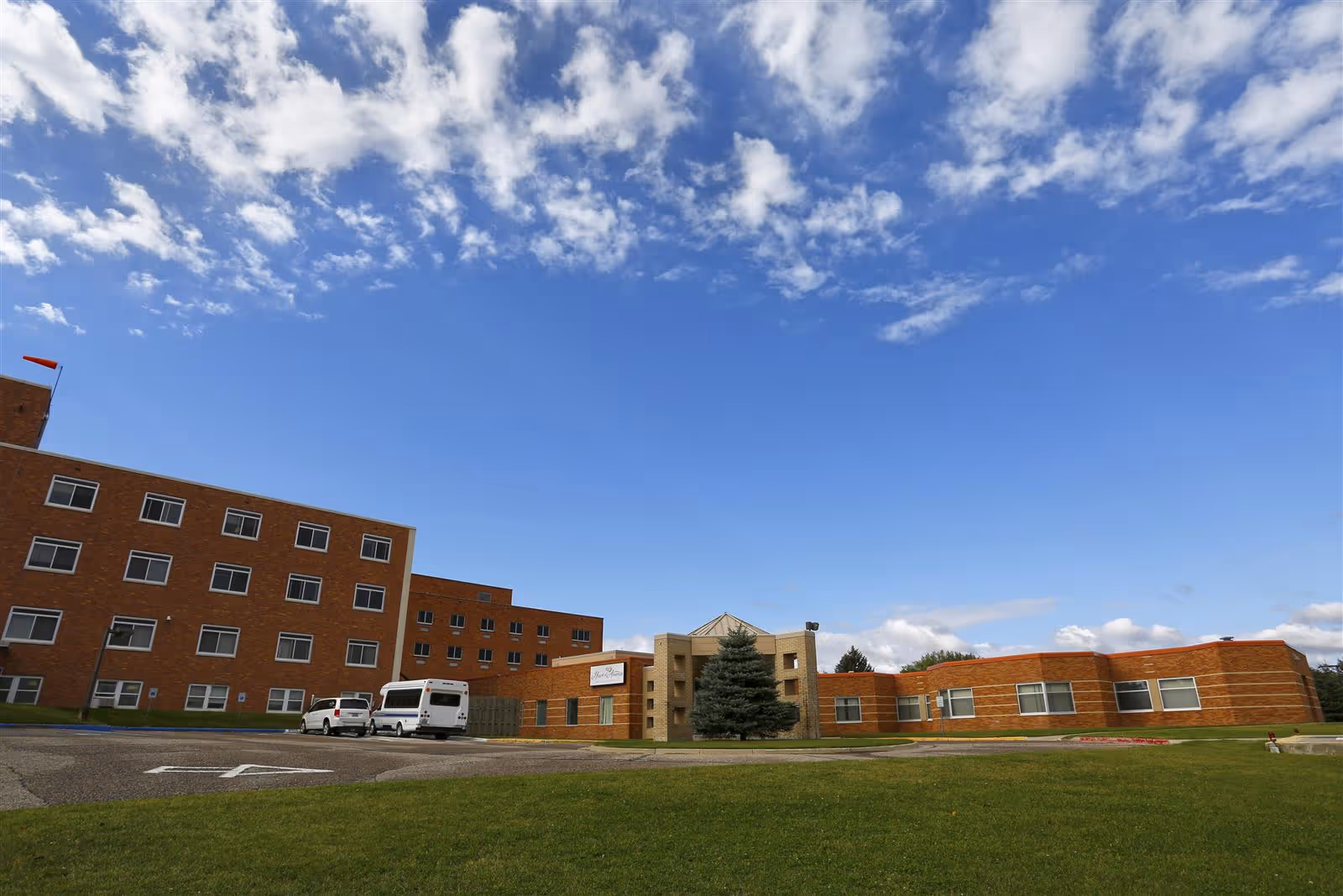 Exterior view of a multi-story brick building under a blue sky with scattered clouds. The building has multiple windows and a small parking area with a few vehicles parked. There is a green lawn in the foreground and a tree near the entrance.