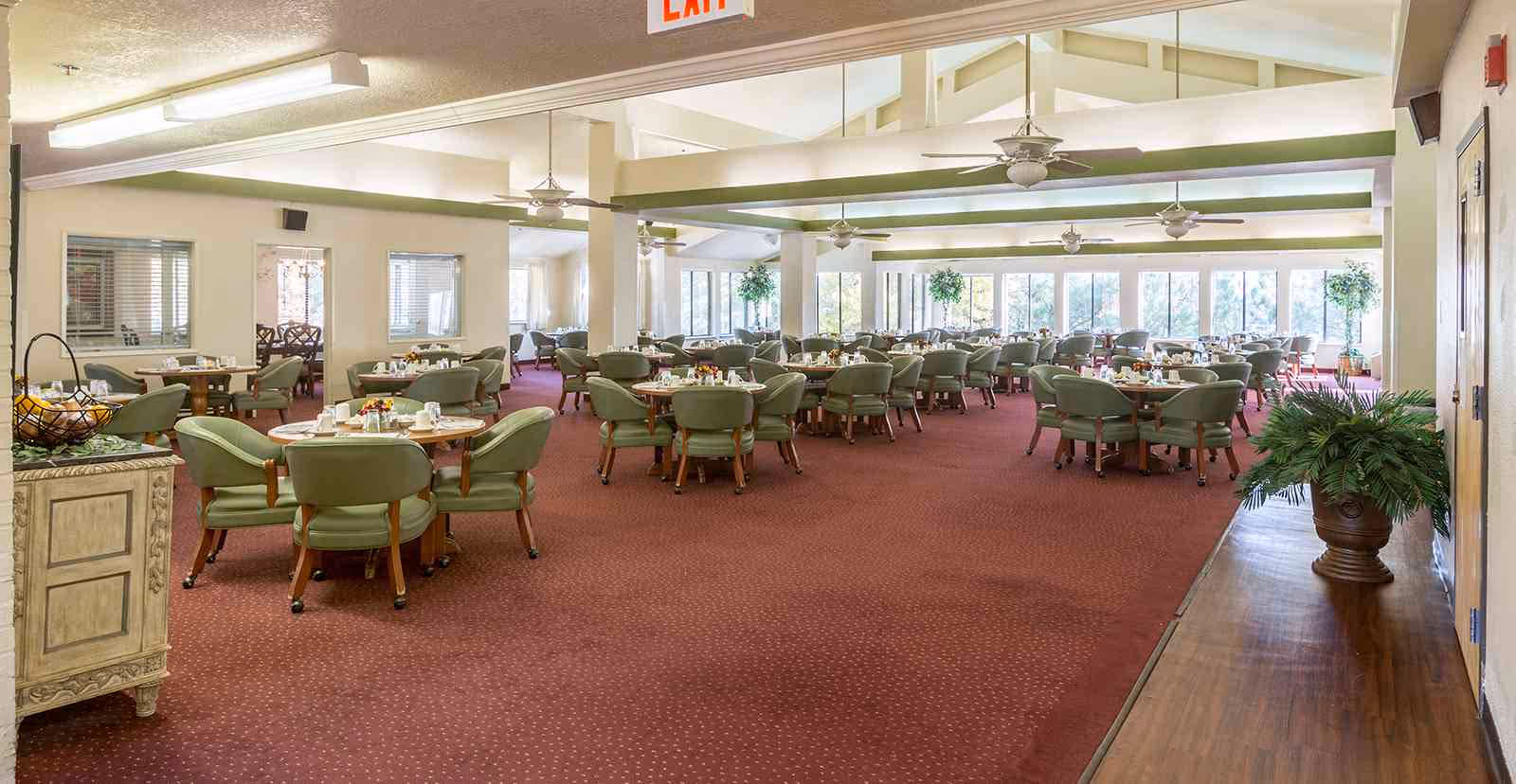 Spacious dining room with multiple round tables set with plates, cups, and condiments. The room has green cushioned chairs, large windows letting in natural light, ceiling fans, and a red carpeted floor. There are plants and decorative elements along the walls.