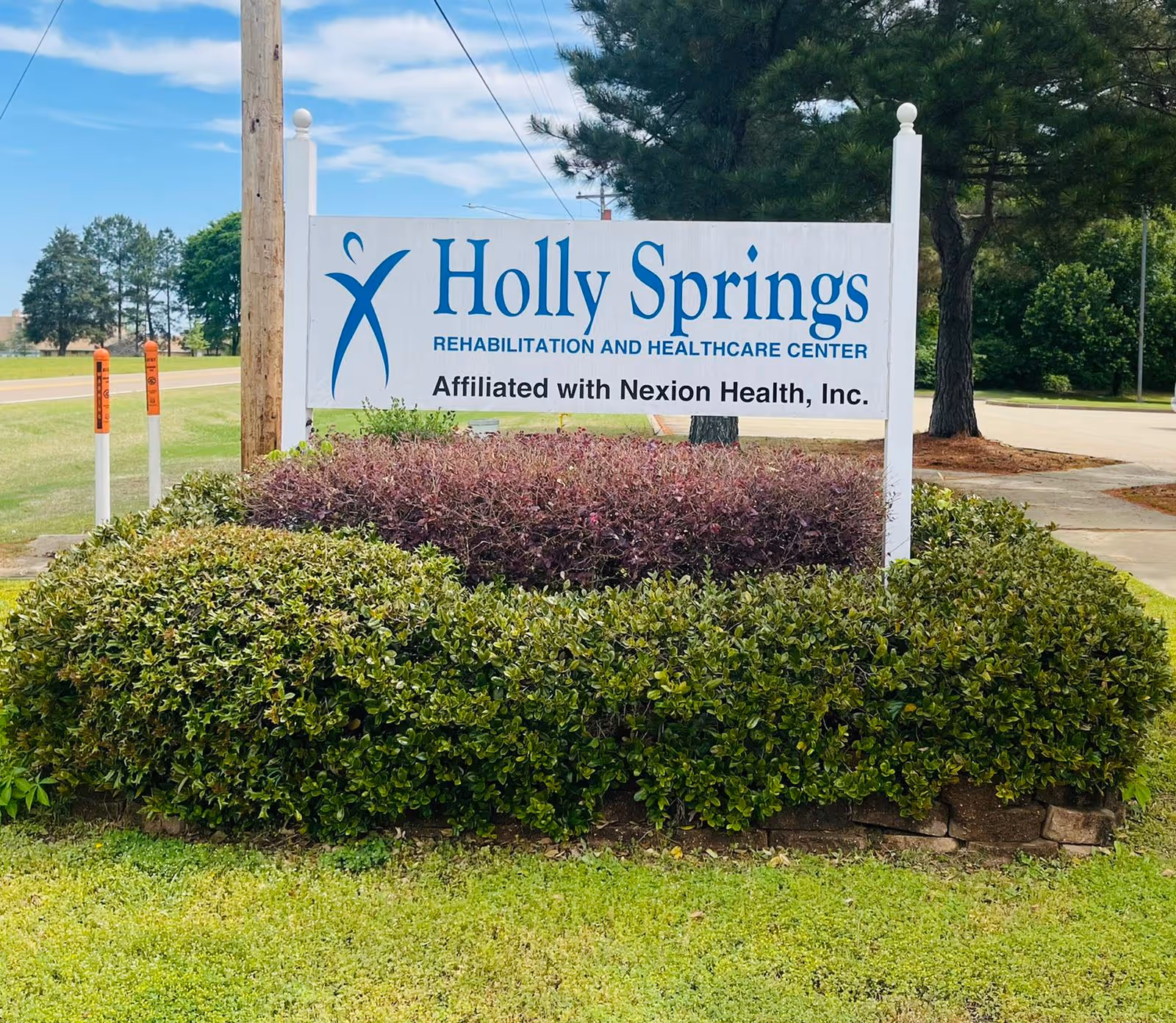 Outdoor view of a white sign for Holly Springs Rehabilitation and Healthcare Center, affiliated with Nexion Health, Inc., surrounded by green bushes and trees with a blue sky in the background.